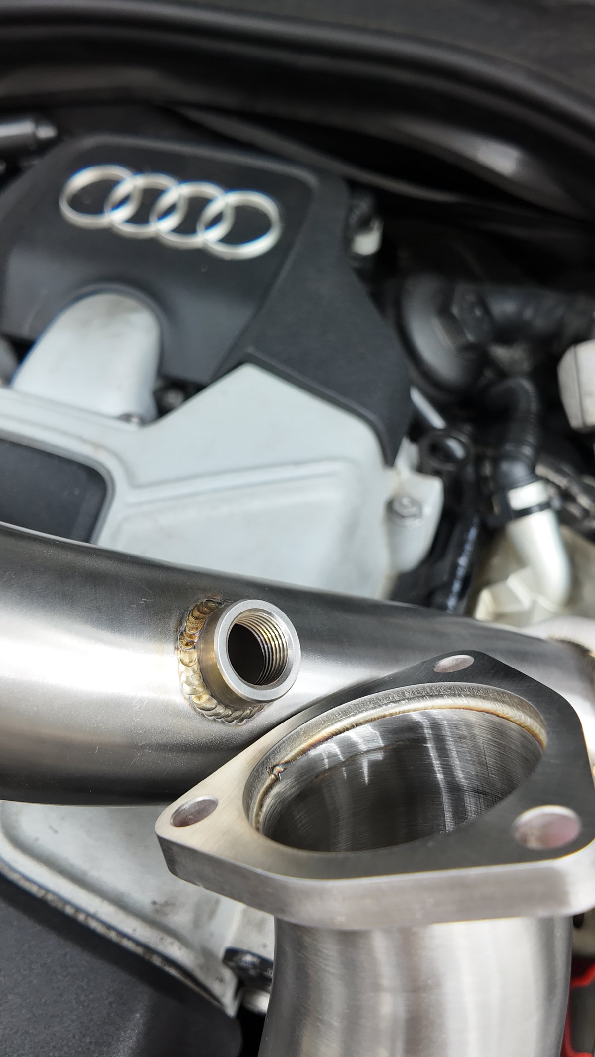 A close-up of a polished stainless steel exhaust pipe with a threaded port and a triangular flange, positioned in front of an Audi engine bay.